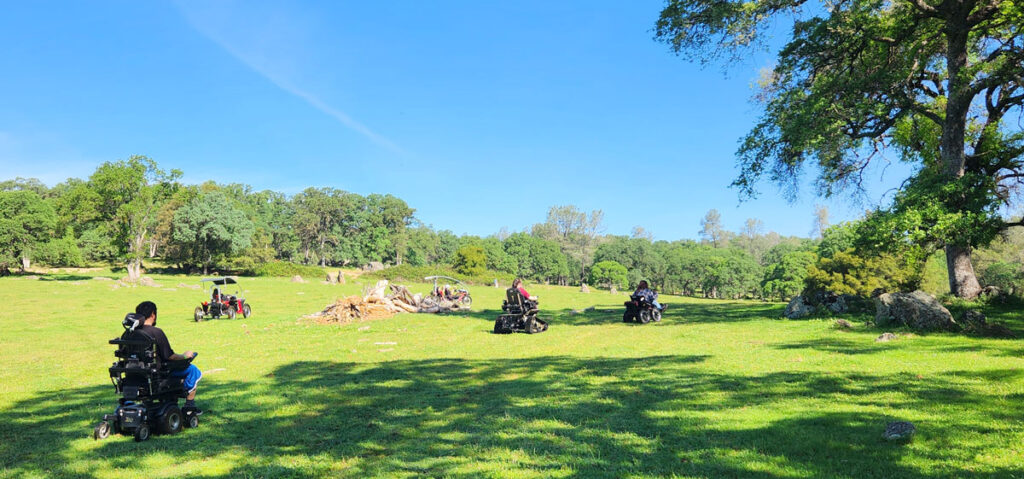Four people use different types of mobility devices to cross the grassy meadow