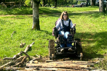 A woman uses a motorized chair to navigate a log crossing over a creek