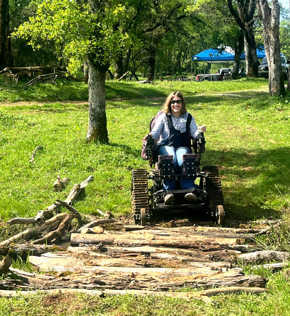 A woman uses a motorized chair to navigate a log crossing over a creek
